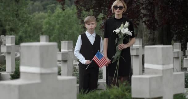 Ritratto di bambino con bandiera americana e giovane donna con fiore che guarda alla macchina fotografica. Donna in abiti scuri che tiene per mano il figlio mentre si trova al cimitero. Concetto di giorno commemorativo. — Video Stock