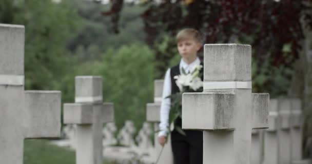 Junge Zicklein mit weißen Lilienblüten auf dem Friedhof. Teenager im Anzug läuft und bleibt in der Nähe des Grabsteins seines Vaters stehen. — Stockvideo