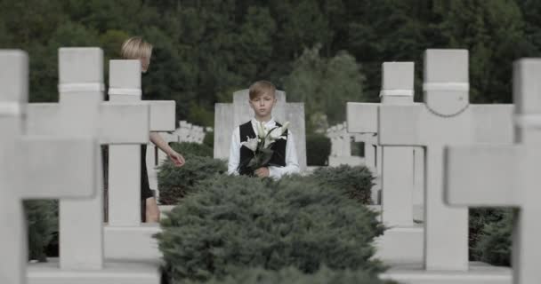 Niño sosteniendo una flor de lirio blanco en el cementerio mientras su madre viene y lo abraza.Niño adolescente mirando a la cámara mientras está de pie en la fila de cruces de piedra. Concepto de día conmemorativo . — Vídeos de Stock