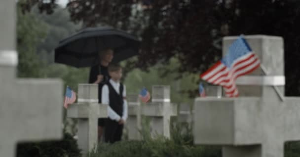 Witwe und ihr halbwüchsiger Sohn stehen unter einem Regenschirm auf dem Friedhof. Mutter und kleiner Junge in der Nähe des Grabsteins. Konzept des Gedenktages. Amerikanische Flaggen auf Steinkreuzen. — Stockvideo