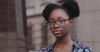 Gözlüklü Afro-Amerikan bir kadının başını çevirip kameraya baktığı yakın plan görüntüsü. Şehir sokaklarında duran ciddi bir milenyum kızının portresi. İnsanlar ve duygular kavramı..