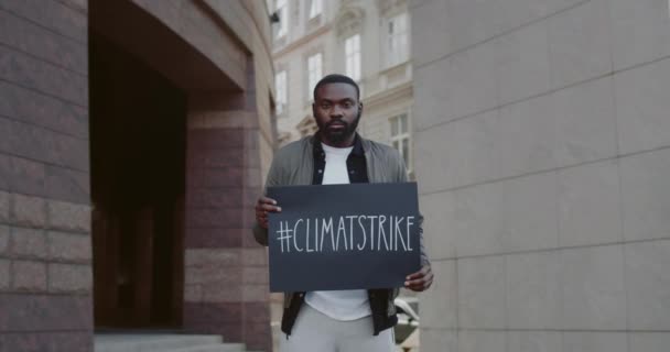 Afro amerikansk man håller plakat med klimatstrejk hashtag när du står på stadens gata. Millennial kille strejkar för ekologi och ren planet. Begreppet rädda jorden. Zooma in. — Stockvideo