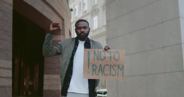 Hombre afroamericano alzando el puño apretado y sosteniendo pancarta de cartón sin racismo. Tipo que apoya el movimiento igualitario de derechos humanos mientras está parado en la calle de la ciudad. . — Vídeos de Stock