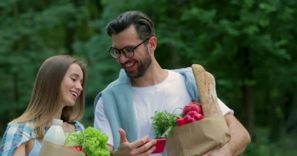 Vista de cerca del hombre y la mujer viendo videos divertidos y hablando mientras llevan bolsas con comida. Pareja alegre sonriendo y mirando la pantalla horizontal del teléfono mientras camina en el parque . — Vídeos de Stock