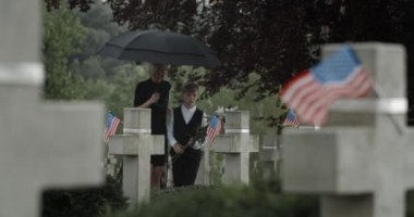 Anne ve küçük oğlu mezarlığa çiçek koyuyorlar. Dul ve çocuk mezar taşının yanında yürüyor ve duruyor. Anma Günü kavramı. Taştan haçlarda Amerikan bayrakları.