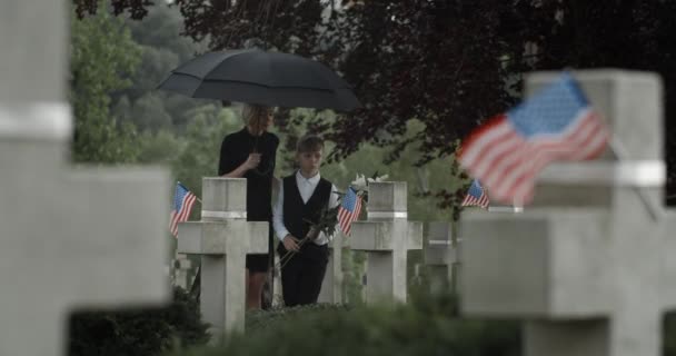 Mother and her teen son under umbrella putting flowers at cemetery. Widow and child walking and stopping near gravestone. Concept of memorial day. American flags on stone crosses. — Stock Video