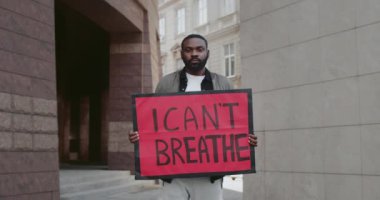 Afro-Amerikalı genç adam, elinde yazılı bir afiş tutuyor. Erkek protestocu sokakta dikilirken polis şiddeti ve ırkçılığına karşı hareketi destekliyor. Yakınlaştır.