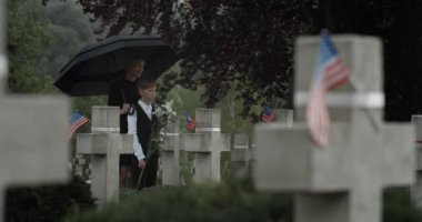 Genç çocuk Kara Dul ve onun mezarlıkta şemsiye altında durması. Anne ve genç çocuk mezar taşının yanında. Anma Günü kavramı. Taştan haçlarda Amerikan bayrakları.