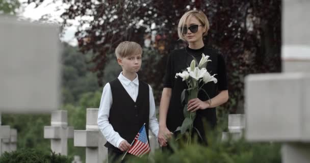 Joven viuda sosteniendo flor y niño con bandera americana mirando el uno al otro y a la cámara. Mujer con ropa oscura sosteniendo a niño de la mano mientras está de pie en el cementerio. Concepto de día conmemorativo. — Vídeos de Stock