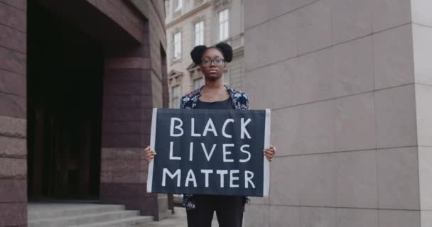 검은 인생을 담은 카튼 플래카드를 들고 있는 안경을 쓴 여학생은 글쓰기가 중요 합니다. 젊은 아프리카계 미국인 여성이 거리에서 서 인종 차별에 반대하는 운동을 지지 했습니다. 줌인. — 비디오