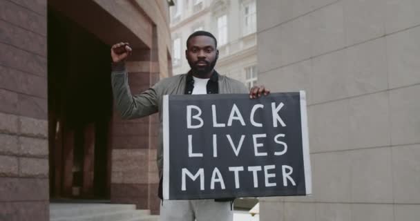 Jóképű szakállas fickó kartondobozban, fekete életrajzokkal. A fiatal afro-amerikai férfi támogatja a rasszizmus elleni mozgalmat, miközben az utcán áll. Közelíts rá!. — Stock videók