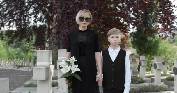 Portrait of widow with flower and young boy looking to camera. Woman in dark clothes and sunglasses holding her teen son hand while they standing at cemetery. Concept of memorial day. — Stock Video