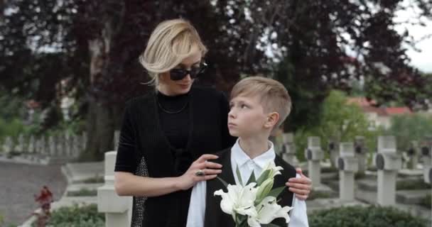 戴太阳镜的年轻寡妇和她的儿子面面相觑，而不是拍照。穿着深色衣服的女人和开着白花的小男孩站在墓地旁.追悼日的概念. — 图库视频影像
