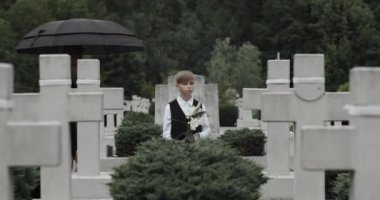 Yas tutan elbiseli bir anne, mezarlıkta çocuğunu çiçeklerle kucaklıyor. Genç bir oğlan ve üzgün genç bir dul şemsiye altında sıra sıra dizilmiş çarmıhlarda dikiliyor. Anma Günü kavramı.