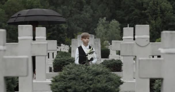 Matka v truchlící oblečení objímající své dítě s květinou na hřbitově.Teen chlapec a smutná mladá vdova stojící v řadě kamenných křížů pod deštníkem. Koncept památného dne. — Stock video