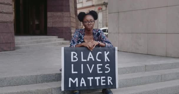 Afrikai-amerikai komoly lány, aki a kamerába néz, miközben a lépcsőnél ül. Egy fiatal nő kartondobozt tart fekete életrajzokkal. A társadalmi probléma fogalma. — Stock videók