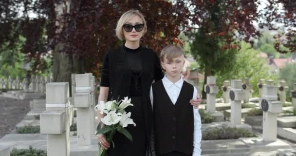 Ung kvinna i svarta kläder som håller blomma och omfamnar sin tonårige son. Änkan i mörka glasögon kramar pojken och tittar på kameran när han står på kyrkogården. Minnesdag. — Stockvideo