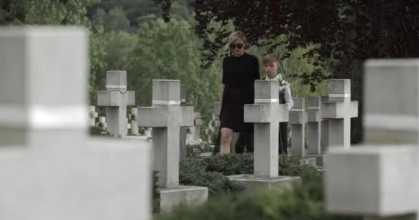 Junge Witwe und ihr Sohn mit weißen Lilienblüten auf dem Friedhof. Teenie-Kind und seine Mutter in dunkler Brille halten Händchen, gehen und halten in der Nähe des Grabsteins seines Vaters.. — Stockvideo