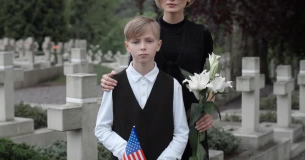 Retrato de mulher em óculos escuros com flor e seu filho olhando para câmera. Jovem segurando bandeira americana enquanto estava com sua mãe no cemitério. Conceito de dia comemorativo . — Vídeo de Stock