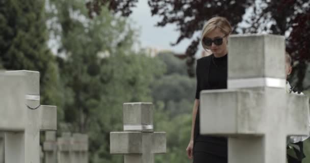 Pohled na matku a jejího dospívajícího syna, jak dávají bílou lilii na hřbitov. Vdova a její dítě se drží za ruce, chodí a zastavují se u náhrobku.. — Stock video
