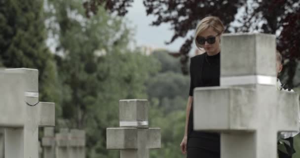 Vista de la cosecha de la madre y su hijo adolescente poniendo flores de lirio blanco en el cementerio. Viuda y su hijo tomados de la mano, caminando y parando cerca de la lápida Concepto de día conmemorativo. — Vídeos de Stock