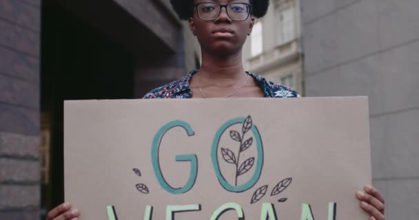 Close-up vista de estudante do sexo feminino segurando Go vegan papelão enquanto está de pé na rua. Afro-americana em óculos promovendo vegetarianismo e estilo de vida saudável . — Vídeo de Stock