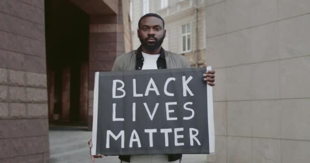 Retrato de un tipo barbudo sosteniendo un cartel de cartón con vidas negras escribiendo materia. Un guapo activista masculino que protesta contra el racismo mientras está parado en la calle. Concepto de igualdad. — Vídeo de stock