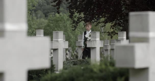 Un ragazzino che viene ad onorare il suo defunto padre al cimitero. Bambino adolescente che cammina e mette fiori di giglio bianchi sulla lapide di suo padre soldato. Concetto di giorno commemorativo. — Video Stock