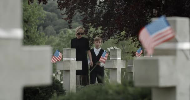 Anne ve küçük oğlu mezarlığa beyaz zambak çiçeği koyuyorlar. Dul ve çocuk el ele tutuşuyor, yürüyor ve mezar taşının yanında duruyor. Anma Günü kavramı. Taştan haçlarda Amerikan bayrakları. — Stok video