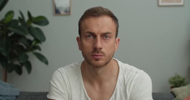 Retrato de un joven guapo mirando a la cámara mientras está sentado en el sofá en la habitación acogedora. Vista de cerca del hombre barbudo serio en los años 30 en chome.Concept de las personas. — Vídeos de Stock