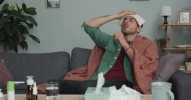 Evdeki kanepede otururken grip ve kötü öksürükten muzdarip genç bir adam. Alnında soğuk kompres tutan 30 'lu yaşlarda sakallı bir adam. Hastalık kavramı.