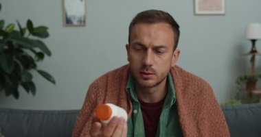 Sakallı genç adam kameraya bakarken konuşuyor ve ilaç gösteriyor. 40 'lı yaşlarda bir adam doktorla internette konuşurken evde kanepede oturuyor..