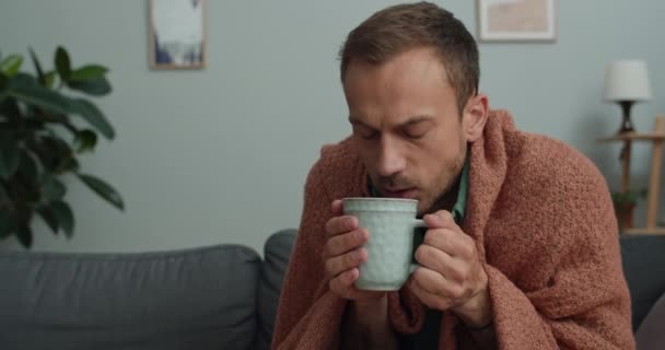 Nahaufnahme von jungen bärtigen Kerl mit Tasse mit heißem Tee und trinken, während sie auf dem Sofa zu Hause sitzen. Kranker Mann um die 40, in Decke gehüllt, wird kalt. Krankheitsbegriff. — Stockvideo