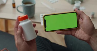 Ekran akıllı telefonunu yatay olarak kurgulayan ve evdeki kanepede otururken plastik hapları kullanan kişinin yakın görüntüsü. Teletıp ve hastalık kavramı.