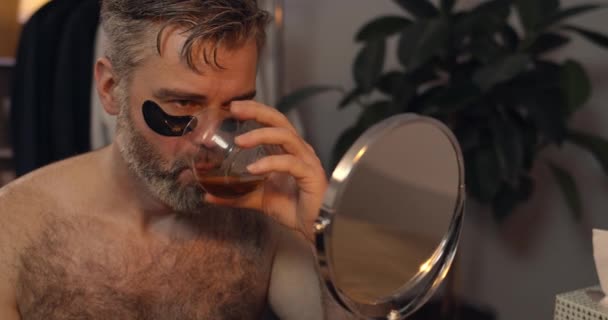 Mature man making painful facial expression while plucking eyebrows in front of mirror.Bearded bachelor with eye patches drinking alcohol before beauty procedure. Home background. — Stock Video