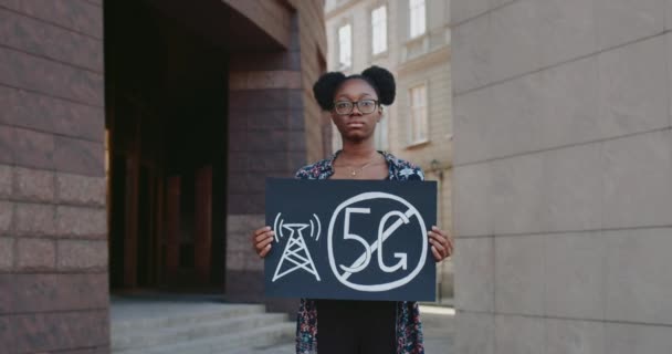 Joven mujer afroamericana sosteniendo pancarta sin letrero de 5g. Estudiante protestando contra la tecnología 5G y el despliegue de antenas compatibles con 5G mientras está parada en la calle. Acercar. — Vídeos de Stock