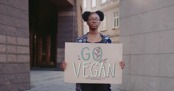 Retrato de una chica afroamericana sosteniendo la pancarta Go Vegan mientras está de pie en la calle.Estudiante femenina con gafas agitando el estilo de vida vegano. Concepto de ir verde y veganismo. — Vídeo de stock