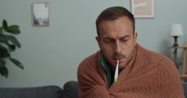 Genç adamın ateşini ölçerken üşüdüğünü yakından görmek. Kanepede otururken battaniyeye sarılı sakallı bir adam termometr 'i ağzında tutuyor..