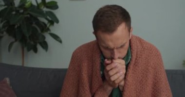 Evdeki kanepede otururken battaniyeye sarılmış genç hasta adam kendini kötü hissediyor. Sakallı adam yüksek sesle öksürüyor ve hastalanırken ürperiyor. Sağlık, hastalık ve grip kavramı.