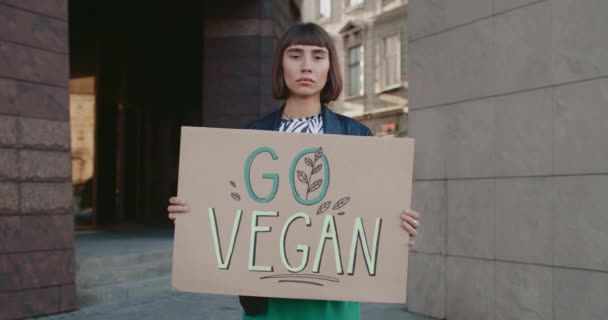 Портрет молодой девушки с плакатом Go Vegan, стоящей на улице. Тысячелетняя женщина с кольцом в носу агитационный вегетарианский образ жизни. Концепция зеленого и веганства. — стоковое видео