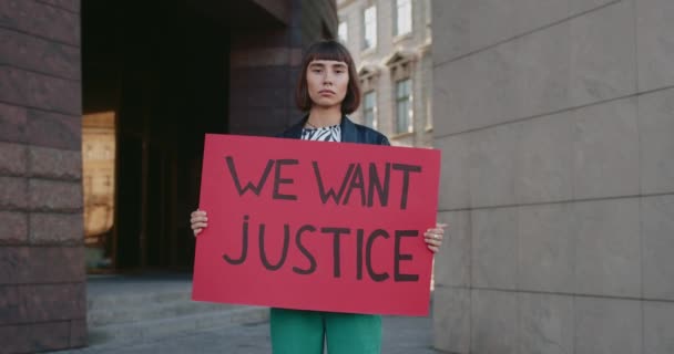 Millennial woman holding carton banner with we want justice writing on it. Hipster girl with nose ring supporting human rights movement while standing at empty street. Zoom in. — Stock Video