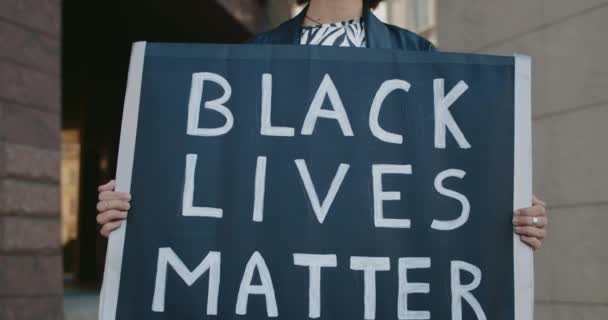 Закройте вид на девушку с картонным плакатом с чёрным жизнью. Жіночий студент, який підтримує рух проти жорстокості поліції та расизму, стоячи на вулиці міста.. — стокове відео