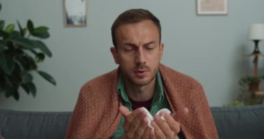 40 'lı yaşlarda hasta bir adam kameraya bakarken hapşırıyor ve burnunu siliyor. Evdeki kanepede otururken internetten doktorla görüşen hasta adam. TeleHealth kavramı.