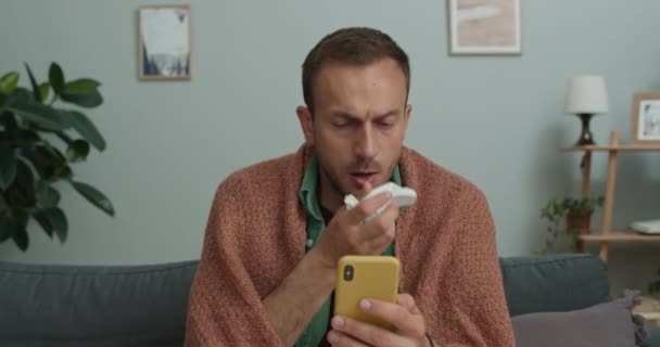 Chlápek ve čtyřiceti používá papírový kapesník, zatímco má videohovor na smartphonu s doktorem. Muž pokrytý přikrývkou s lékařskou konzultaci, zatímco sedí na gauči doma. — Stock video
