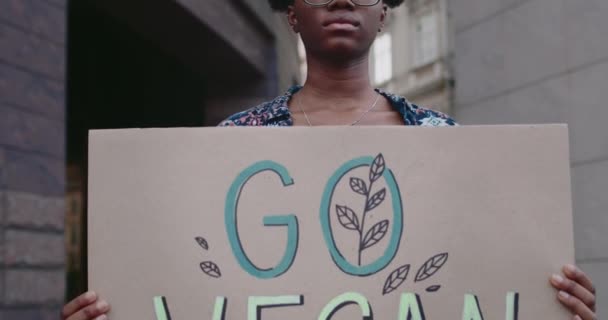 Vista de cerca de la chica afroamericana sosteniendo Go Vegan pancarta mientras está de pie en la calle. Activista femenina en gafas agitando el estilo de vida vegetariano. Concepto de ir verde y veganismo. — Vídeos de Stock