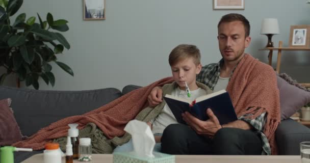 Ojciec czyta książkę i jego chory syn mierzą temperaturę leżąc na sofie. Piękna matka przynosi gorący napój i bierze termometr. Pojęcie choroby i opieki zdrowotnej. — Wideo stockowe