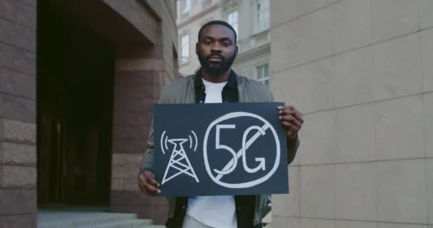 Porträtt av stilig afro amerikan man håller plakat utan 5g tecken. Skäggig kille protesterar mot ny teknik och kompatibel antennutplacering stående på gatan. — Stockvideo