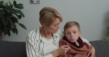 Genç bir kadının doktorla görüntülü konuşma yaparken termometreye bakıp konuşmasına yakından bakın. Anne ve oğlu çevrimiçi tıbbi danışmanlık yaparken kameraya bakıyor..