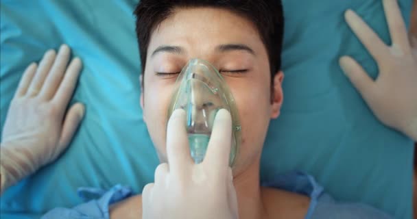 Sluiten van bewusteloze Aziatische man met zuurstof inhalatie masker sterven terwijl liggend op medische brancard. Paramedici controleren pols naar millennial guy terwijl ze naar operatiekamer. — Stockvideo