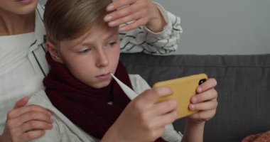 Hasta çocuğun internet üzerinden oyun oynamasını ve sıcaklığı ölçmesini kapat. Anne, oğlunun alnına dokunuyor ve kanepede otururken termometre alıyor..